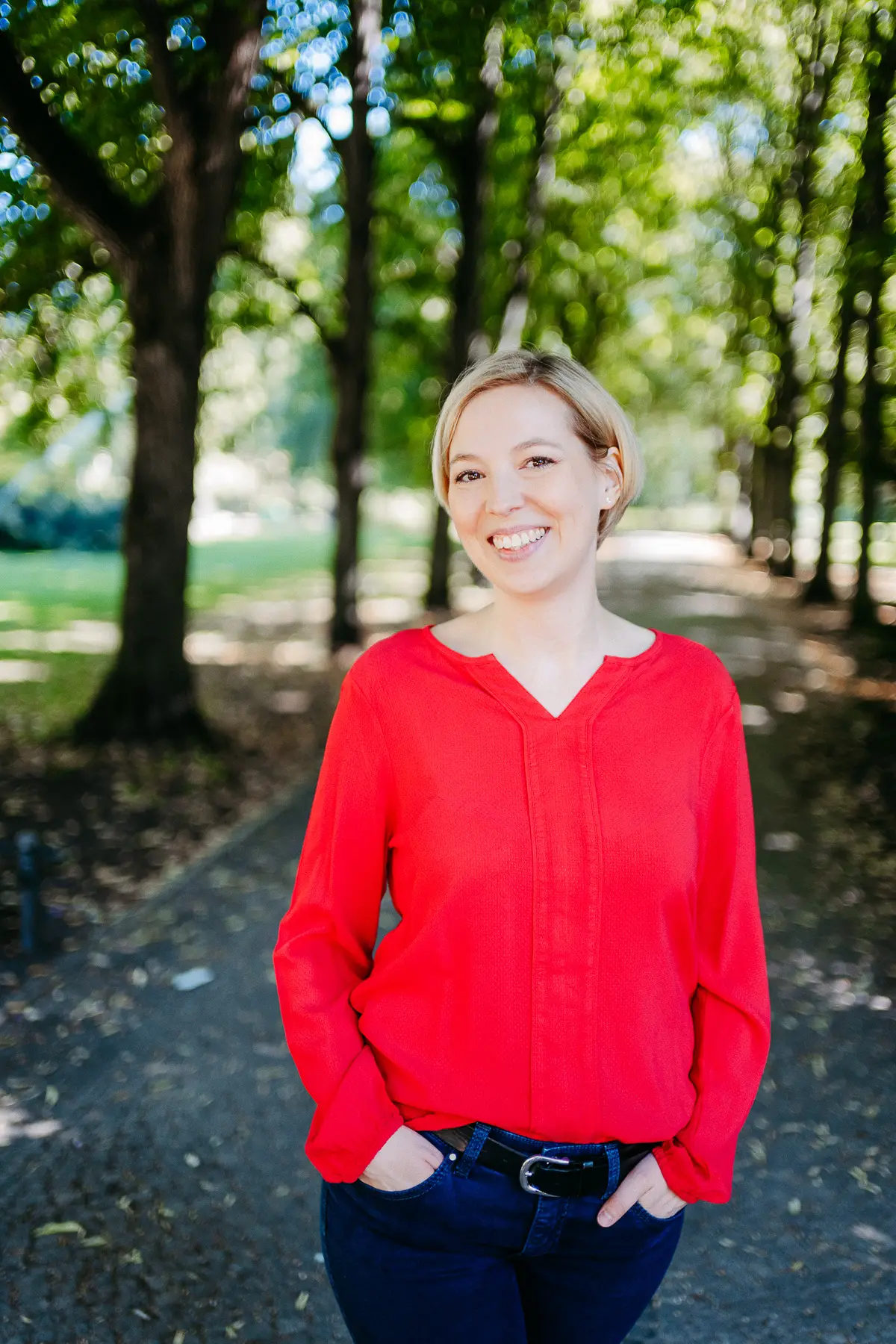 Gute Hochzeitsrednerin steht in roter Bluse in einem schoenen Park zwischen Baeumen